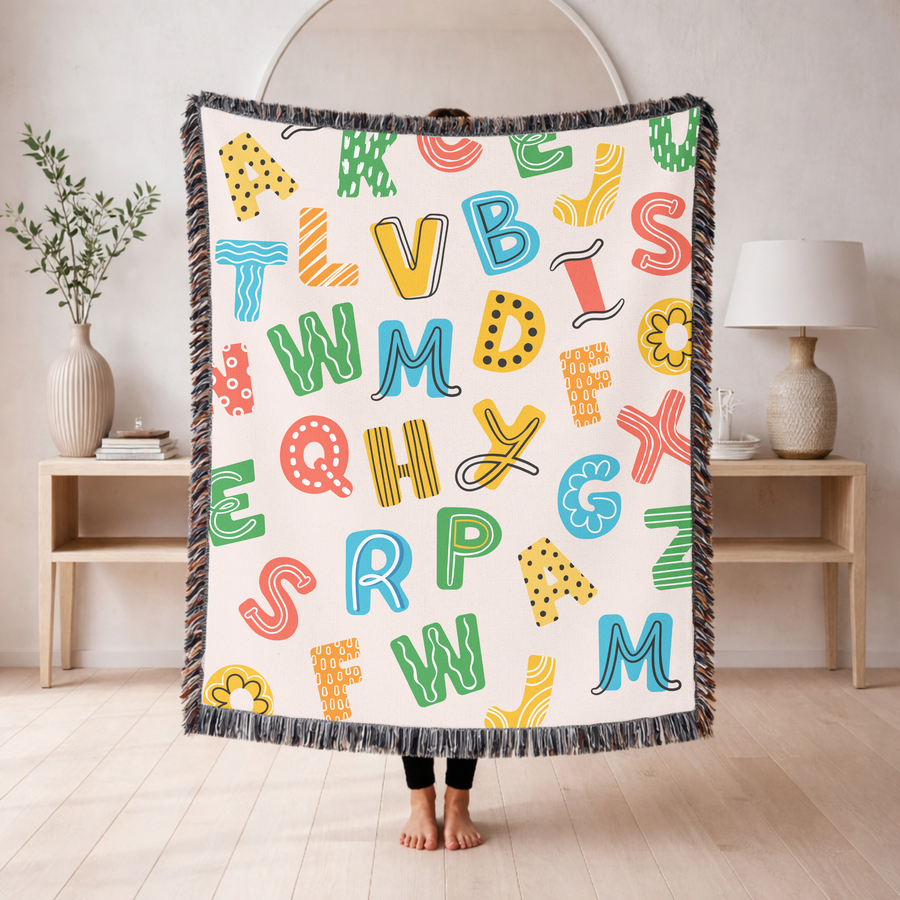 Cream fringed woven blanket scattered with big playful alphabet letters in green, blue, yellow and coral, each filled with dots, stripes or squiggles, with a few flowers between.