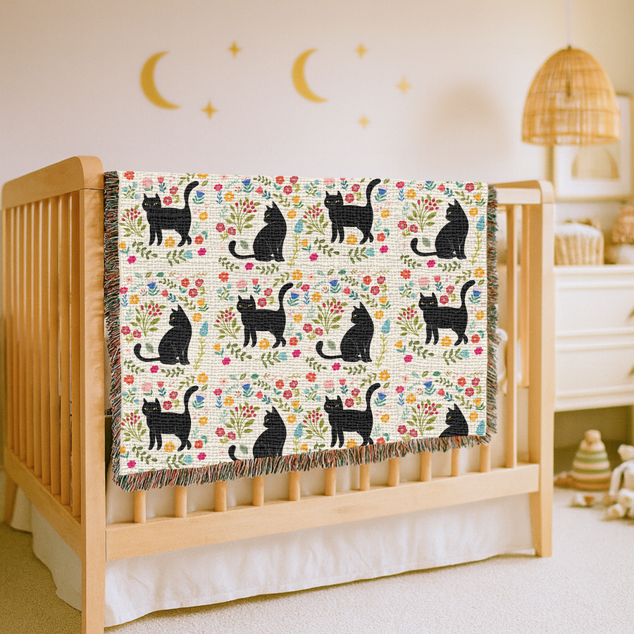 Cream fringed woven blanket patterned with black cats sitting, standing and stretching among small folk-art flowers in pink, yellow, blue and red on curling green leafy stems.