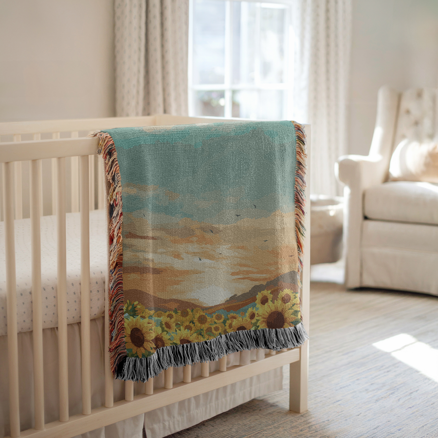 Fringed woven blanket printed with a field of open sunflowers below rolling hills, a wide sunset sky of teal and apricot cloud above them and small birds crossing the light.