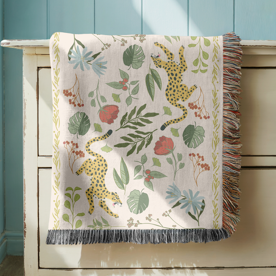 Cream fringed woven blanket printed with two spotted leopards climbing among tropical leaves, their tails curling behind them, with blue daisies, red poppies and orange berries.