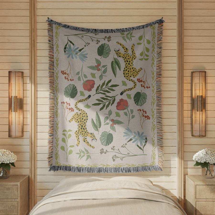 Cream fringed woven blanket printed with two spotted leopards climbing among tropical leaves, their tails curling behind them, with blue daisies, red poppies and orange berries.