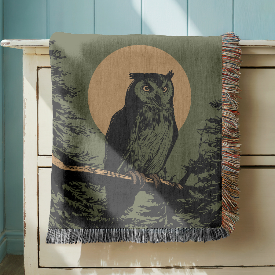 Olive green fringed woven blanket printed with a horned owl perched on a branch against a large cream moon, its eyes lit, with dark pine silhouettes rising on either side.