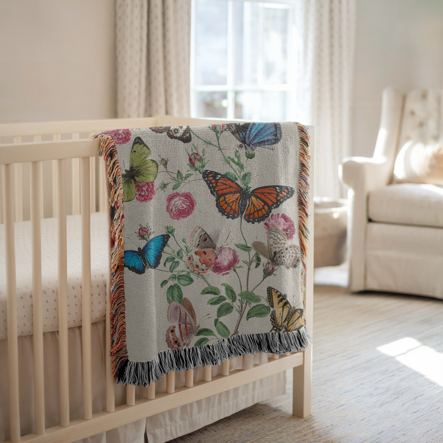 Cream fringed woven blanket printed with botanical butterflies, a blue morpho, a monarch and a swallowtail among them, resting on climbing stems of pink roses and small buds.