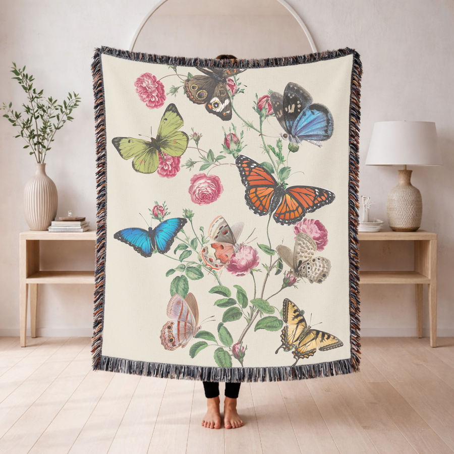 Cream fringed woven blanket printed with botanical butterflies, a blue morpho, a monarch and a swallowtail among them, resting on climbing stems of pink roses and small buds.