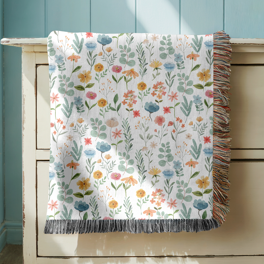 White fringed woven blanket scattered with watercolour wildflowers in blue, yellow, coral and pink, their slender stems, fine grasses and green foliage spread across the cloth.