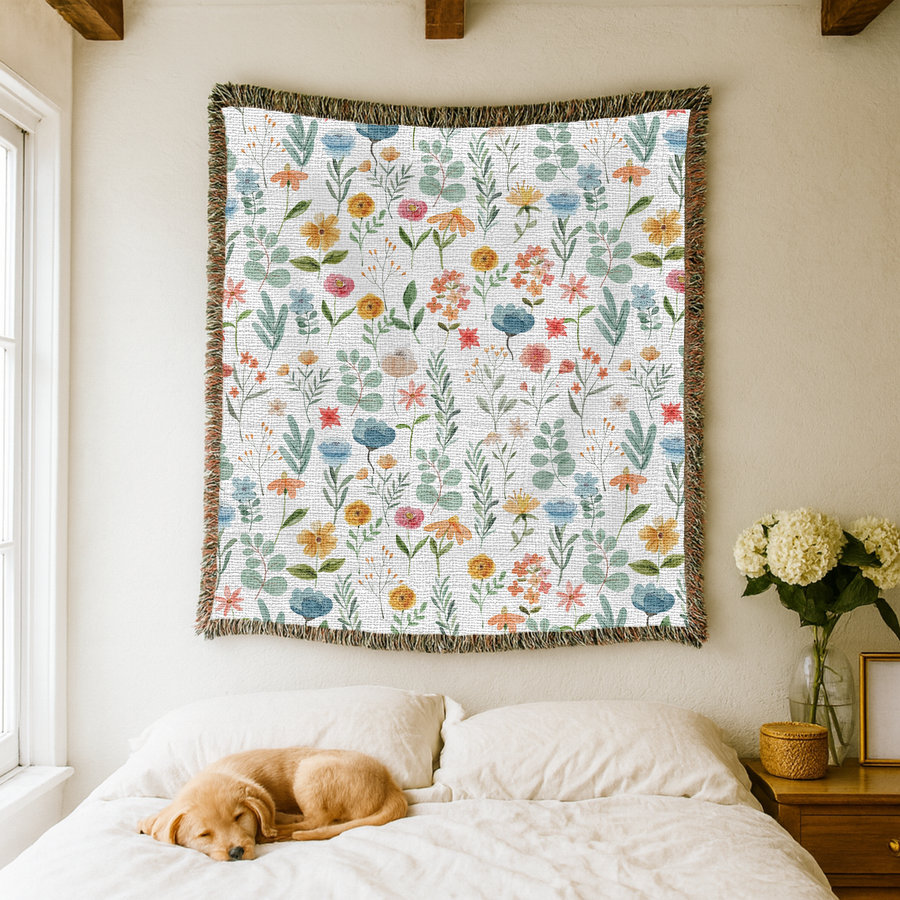 White fringed woven blanket scattered with watercolour wildflowers in blue, yellow, coral and pink, their slender stems, fine grasses and green foliage spread across the cloth.