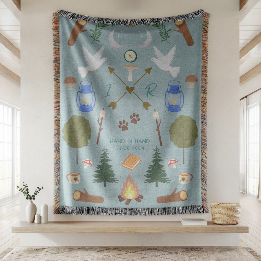 Pale blue fringed woven blanket of camping motifs, doves, lanterns, pines, a campfire and marshmallows, with the initials I and R on crossed arrows above Hand In Hand Since 2004.