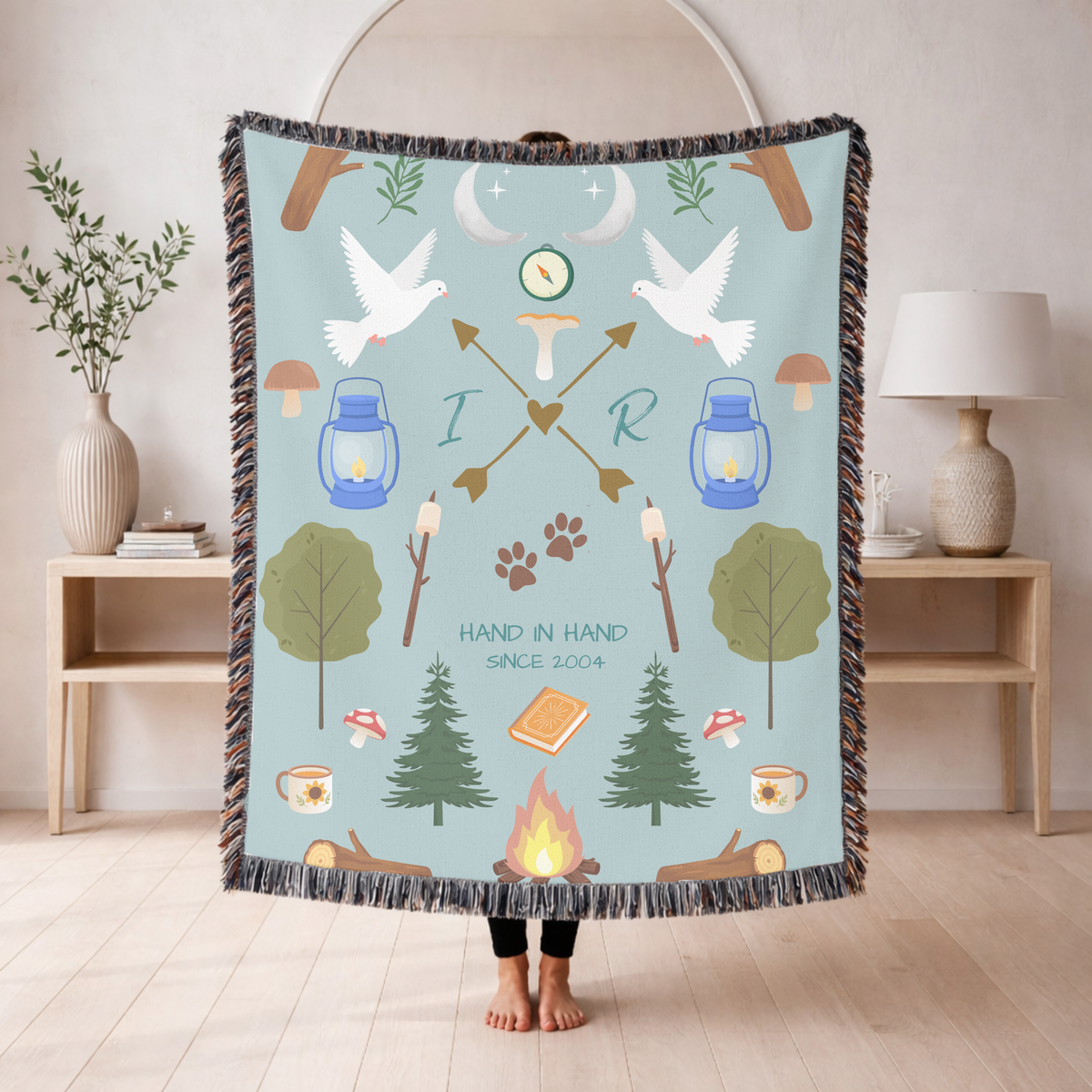 Pale blue fringed woven blanket of camping motifs, doves, lanterns, pines, a campfire and marshmallows, with the initials I and R on crossed arrows above Hand In Hand Since 2004.