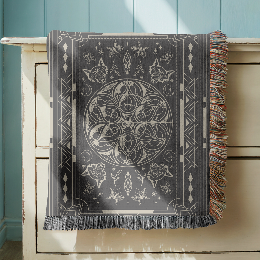 Charcoal fringed woven blanket printed in cream with an ornate bordered panel, a central knot of coiled snakes, wolf heads at the corners, moths, roses and crescent moons.
