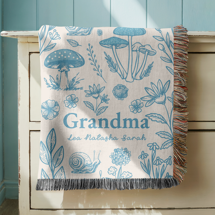 Cream fringed woven blanket printed in blue line work with toadstools, flowers, leaves, a snail and a dandelion clock, reading Grandma above the names Lea, Natasha and Sarah.