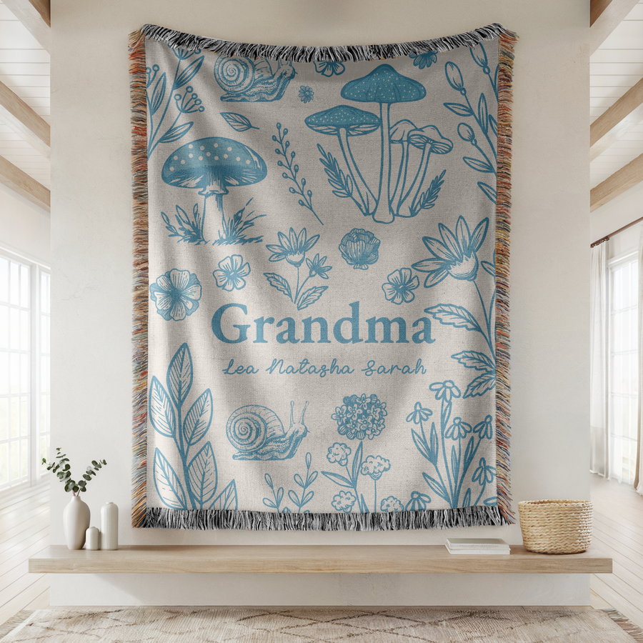 Cream fringed woven blanket printed in blue line work with toadstools, flowers, leaves, a snail and a dandelion clock, reading Grandma above the names Lea, Natasha and Sarah.
