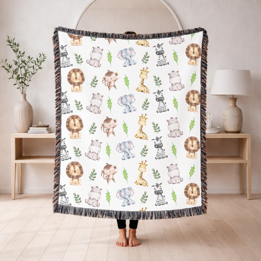 White fringed woven blanket printed with watercolour safari animals, lions, elephants, giraffes, zebras, hippos and monkeys, each drawn in soft washes among small green leaves.