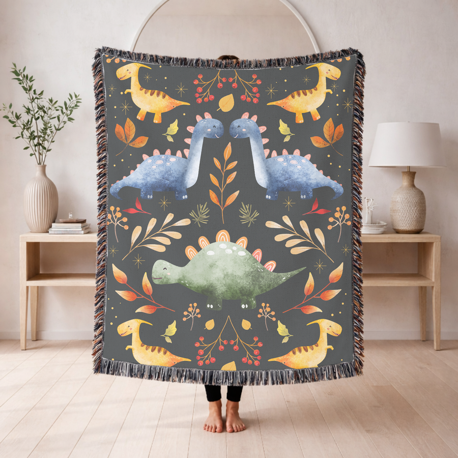 Charcoal fringed woven blanket printed with folk-art dinosaurs in yellow, blue and green, arranged in mirrored pairs among autumn leaves, red berries and small gold stars.