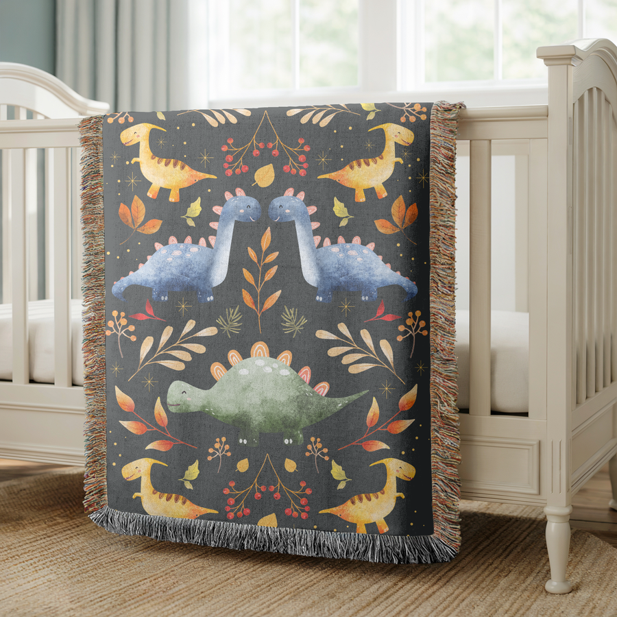 Charcoal fringed woven blanket printed with folk-art dinosaurs in yellow, blue and green, arranged in mirrored pairs among autumn leaves, red berries and small gold stars.