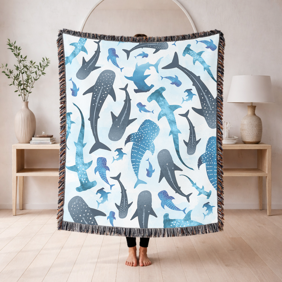Pale blue fringed woven blanket printed with watercolour sharks, whale sharks, hammerheads and smaller fish, some spotted and some plain, drifting across a washed background.