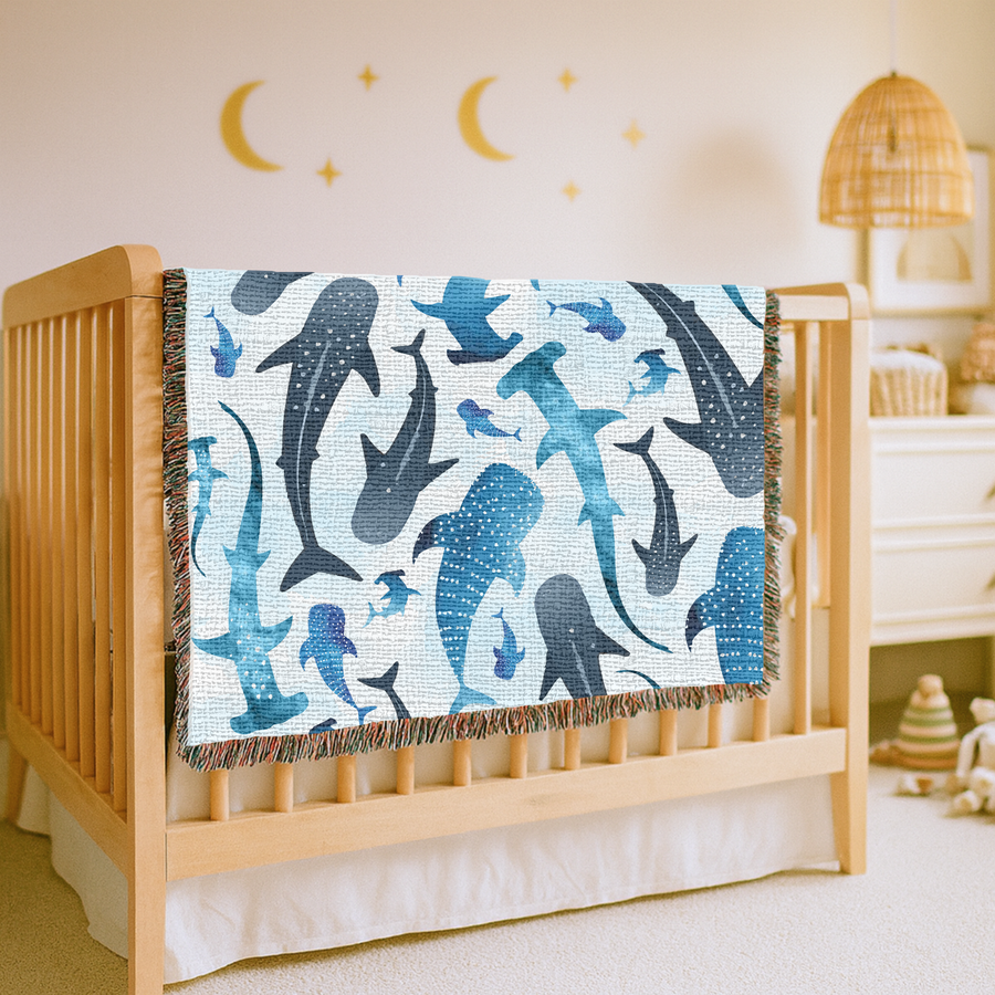 Pale blue fringed woven blanket printed with watercolour sharks, whale sharks, hammerheads and smaller fish, some spotted and some plain, drifting across a washed background.