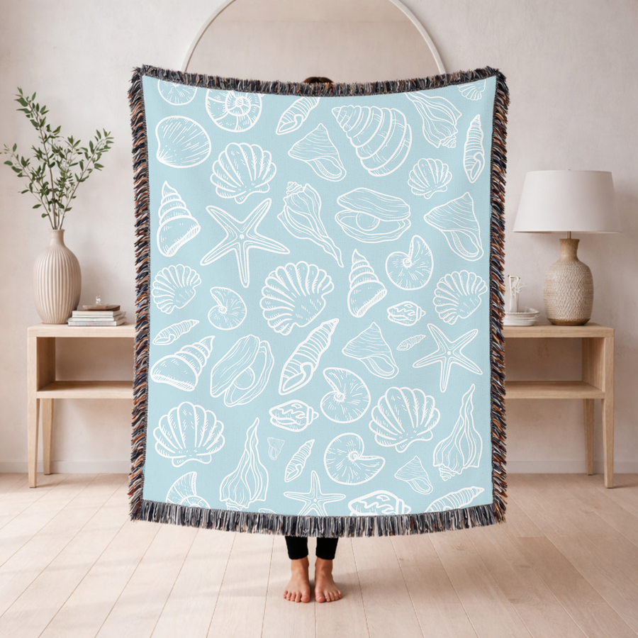 Pale blue fringed woven blanket printed with scallop shells, spirals, starfish and a nautilus drawn as simple white outlines, set in even rows across the whole of the cloth.