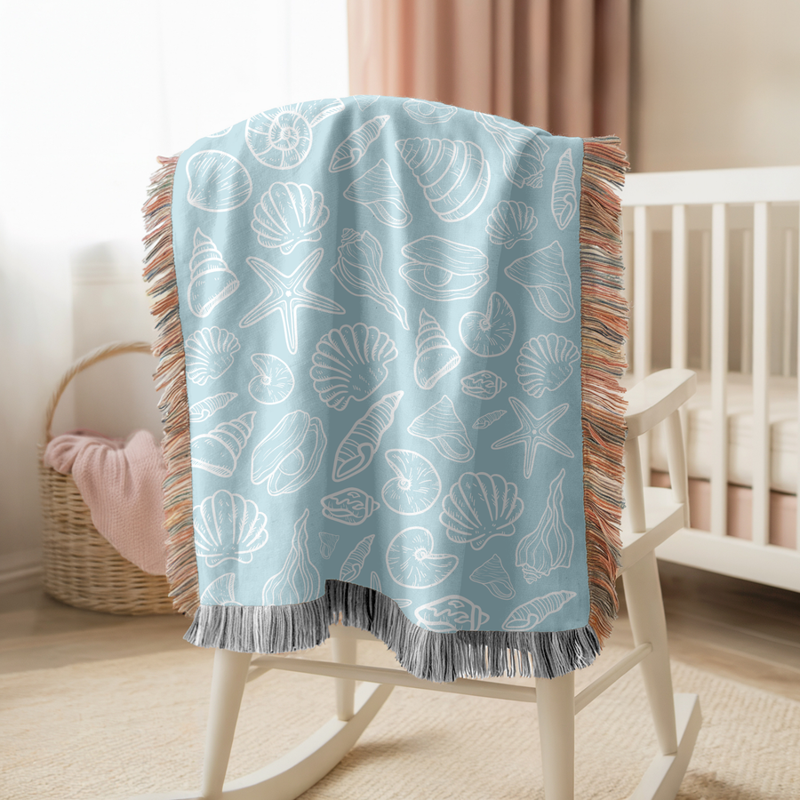 Pale blue fringed woven blanket printed with scallop shells, spirals, starfish and a nautilus drawn as simple white outlines, set in even rows across the whole of the cloth.