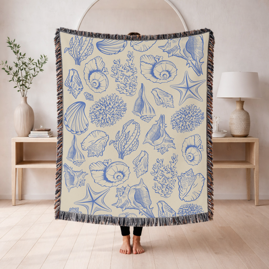 Cream fringed woven blanket printed with shells, coral, a starfish and a nautilus drawn in fine blue line work, arranged loosely across the ground like an old engraved plate.
