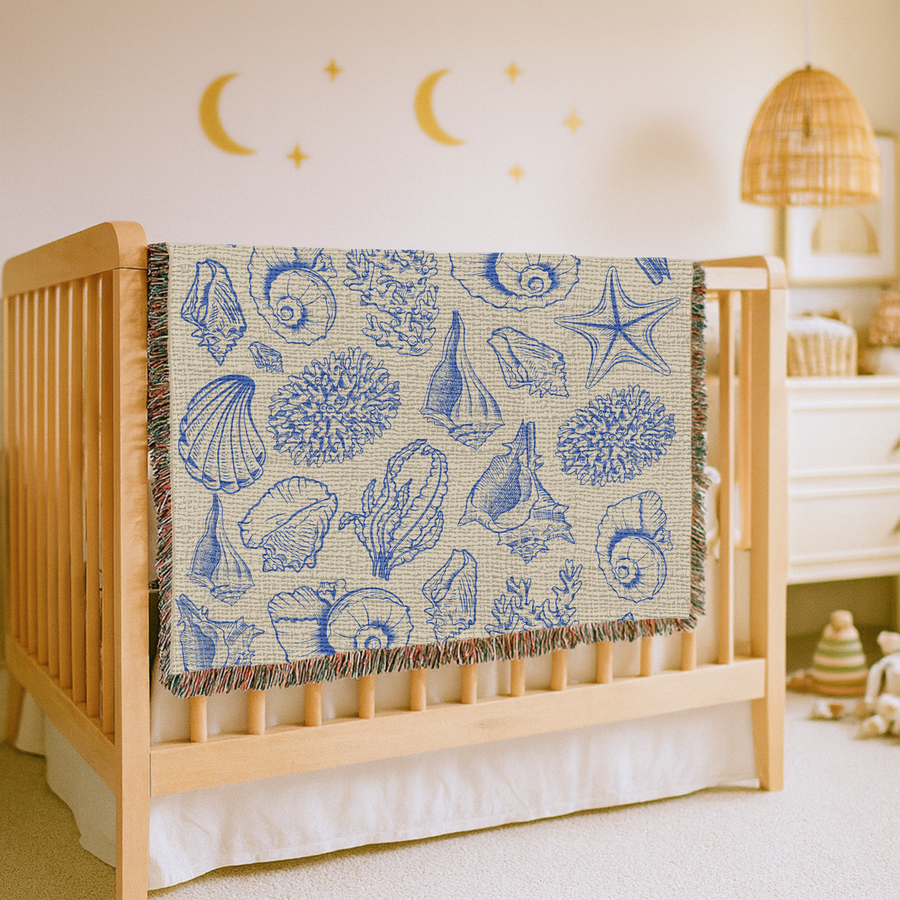 Cream fringed woven blanket printed with shells, coral, a starfish and a nautilus drawn in fine blue line work, arranged loosely across the ground like an old engraved plate.