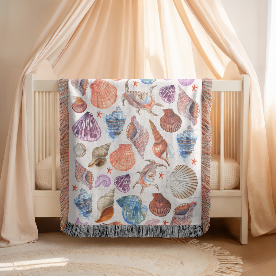 Cream fringed woven blanket printed with watercolour seashells in coral, purple, blue and tan, with small orange starfish, a spiral snail shell and a pearl in an open oyster.
