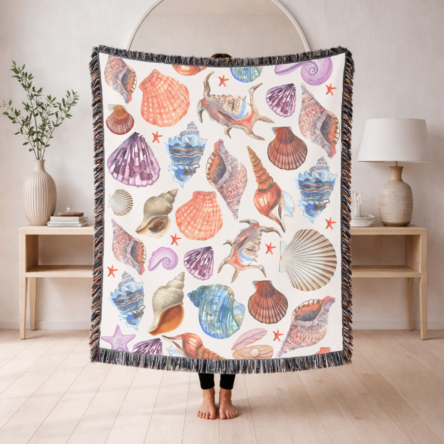 Cream fringed woven blanket printed with watercolour seashells in coral, purple, blue and tan, with small orange starfish, a spiral snail shell and a pearl in an open oyster.