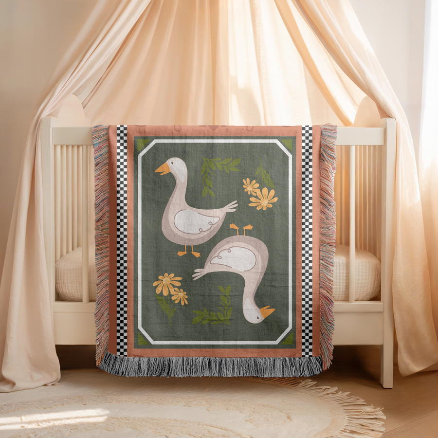 Fringed woven blanket with a terracotta and checkerboard border framing an olive panel, where two cream geese stand among yellow daisies and small sprigs of green leaves.