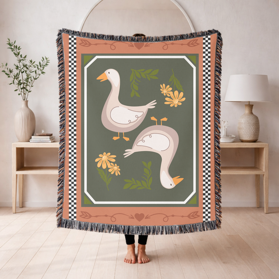 Fringed woven blanket with a terracotta and checkerboard border framing an olive panel, where two cream geese stand among yellow daisies and small sprigs of green leaves.