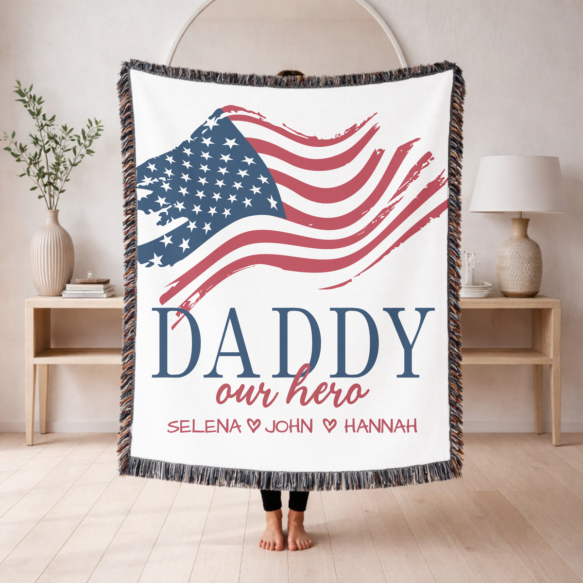 White fringed woven blanket printed with a brushstroke American flag above the word Daddy in navy and our hero in red script, over the names Selena, John and Hannah in red.
