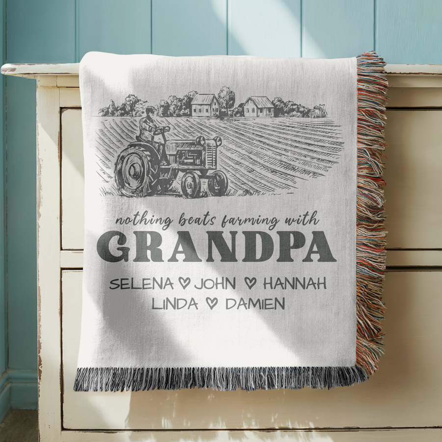 Cream fringed woven blanket with an engraved farm scene of a tractor and farmhouses, reading nothing beats farming with Grandpa above five names divided by small dark hearts.