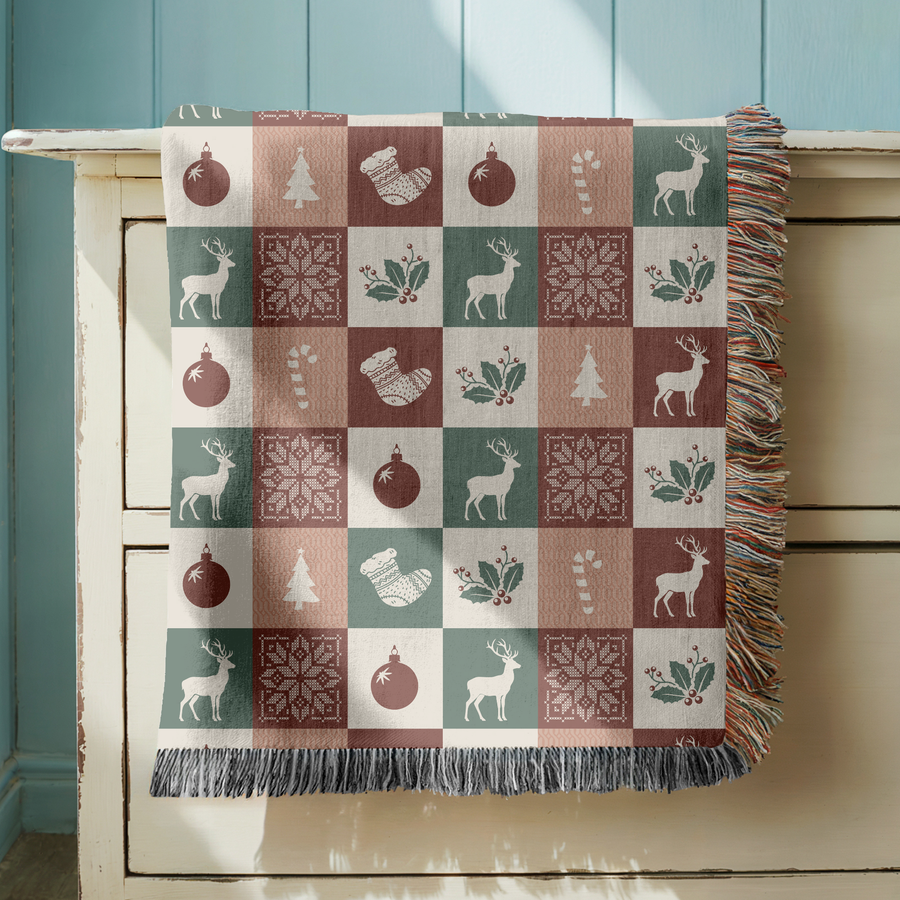 Fringed woven blanket in a checkerboard of green, rose, cream and burgundy squares, each holding a reindeer, a bauble, a stocking, a candy cane, a tree, holly or a snowflake.