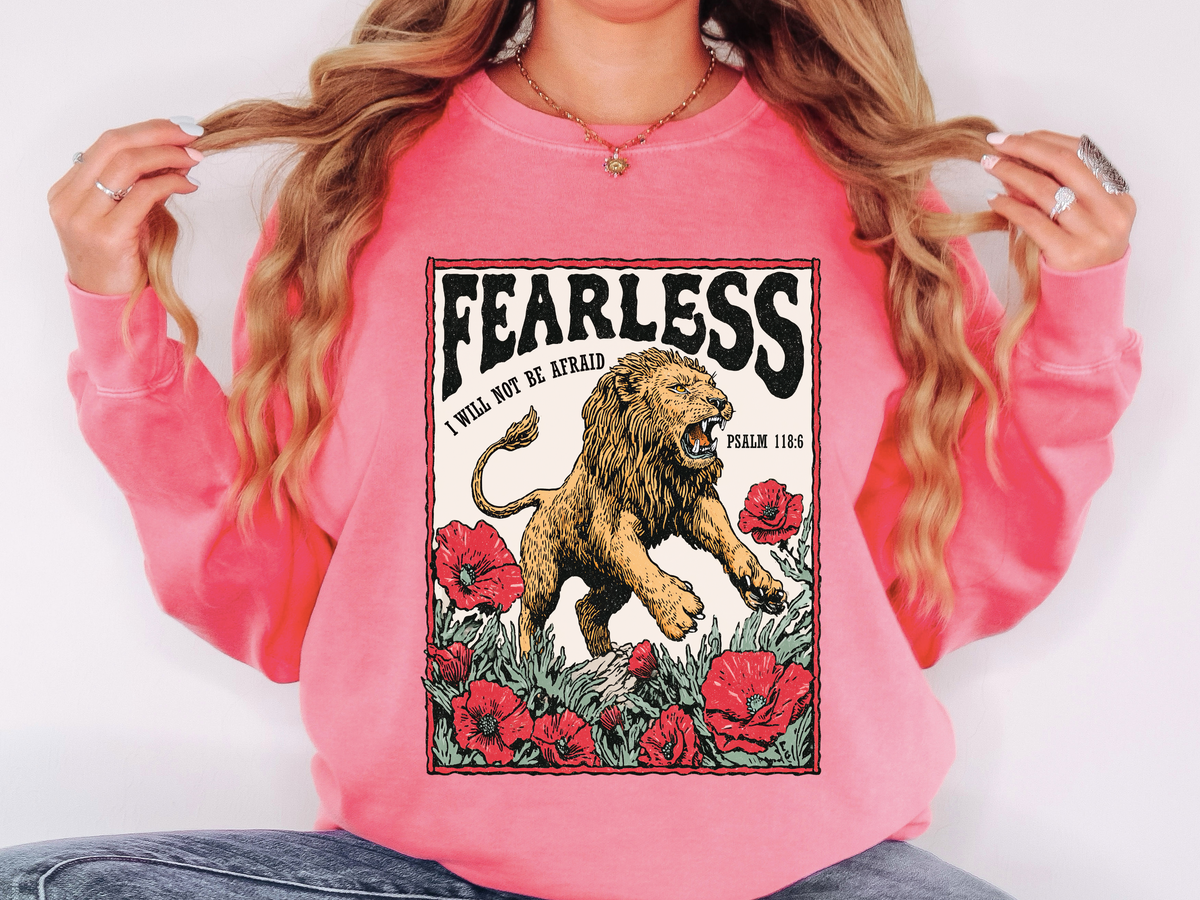 A pink crewneck sweatshirt printed with a roaring lion among red roses under Fearless and I will not be afraid, Psalm 118:6.
