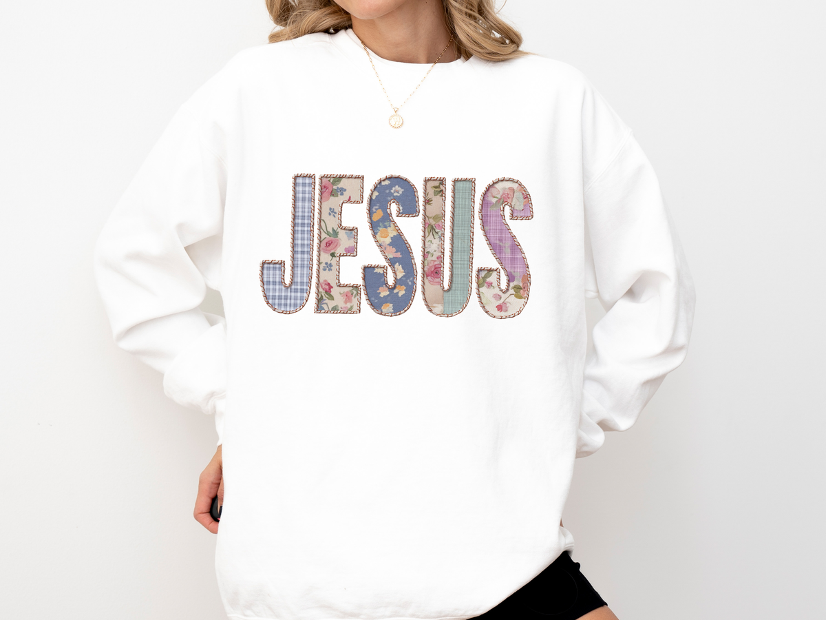 A white crewneck sweatshirt printed with JESUS in patchwork letters of gingham, floral and plaid fabric.