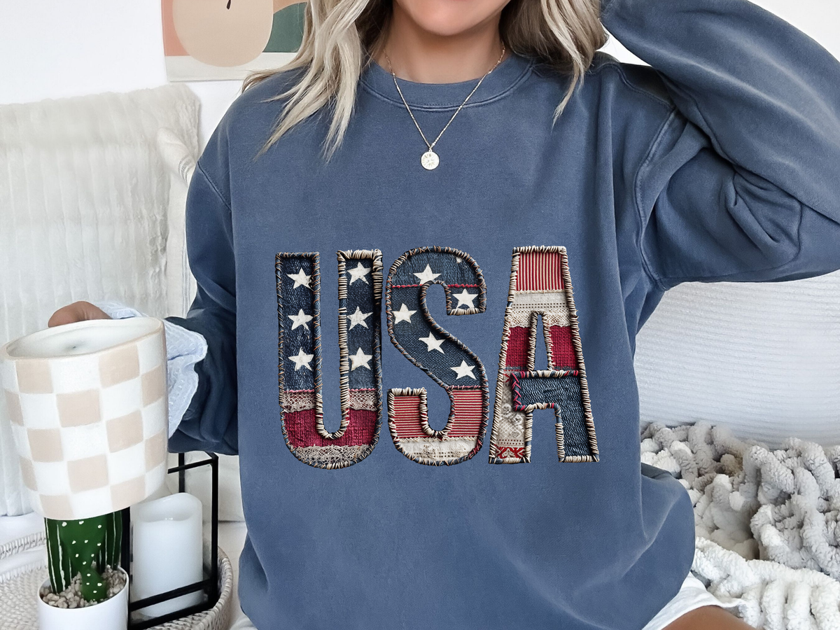 A blue crewneck sweatshirt printed with USA in stitched patchwork letters of stars and stripes.
