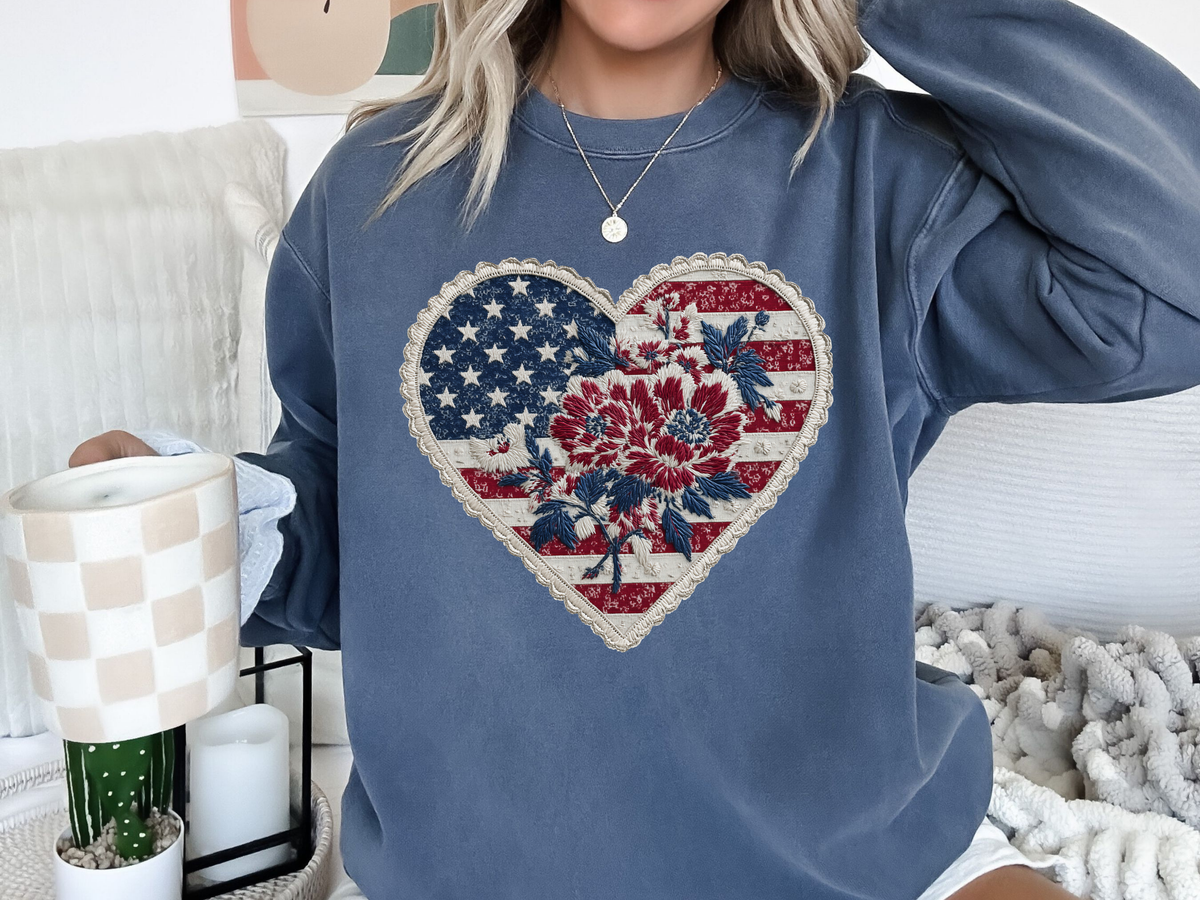 A blue crewneck sweatshirt printed with a heart-shaped stars-and-stripes patch with dark red flowers and a stitched border.