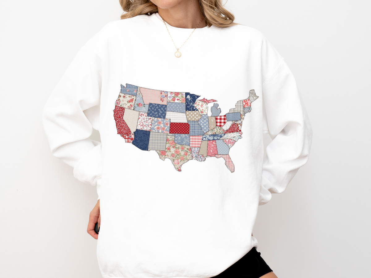 A white crewneck sweatshirt printed with a map of the United States made of patchwork quilt squares in red, blue and cream.