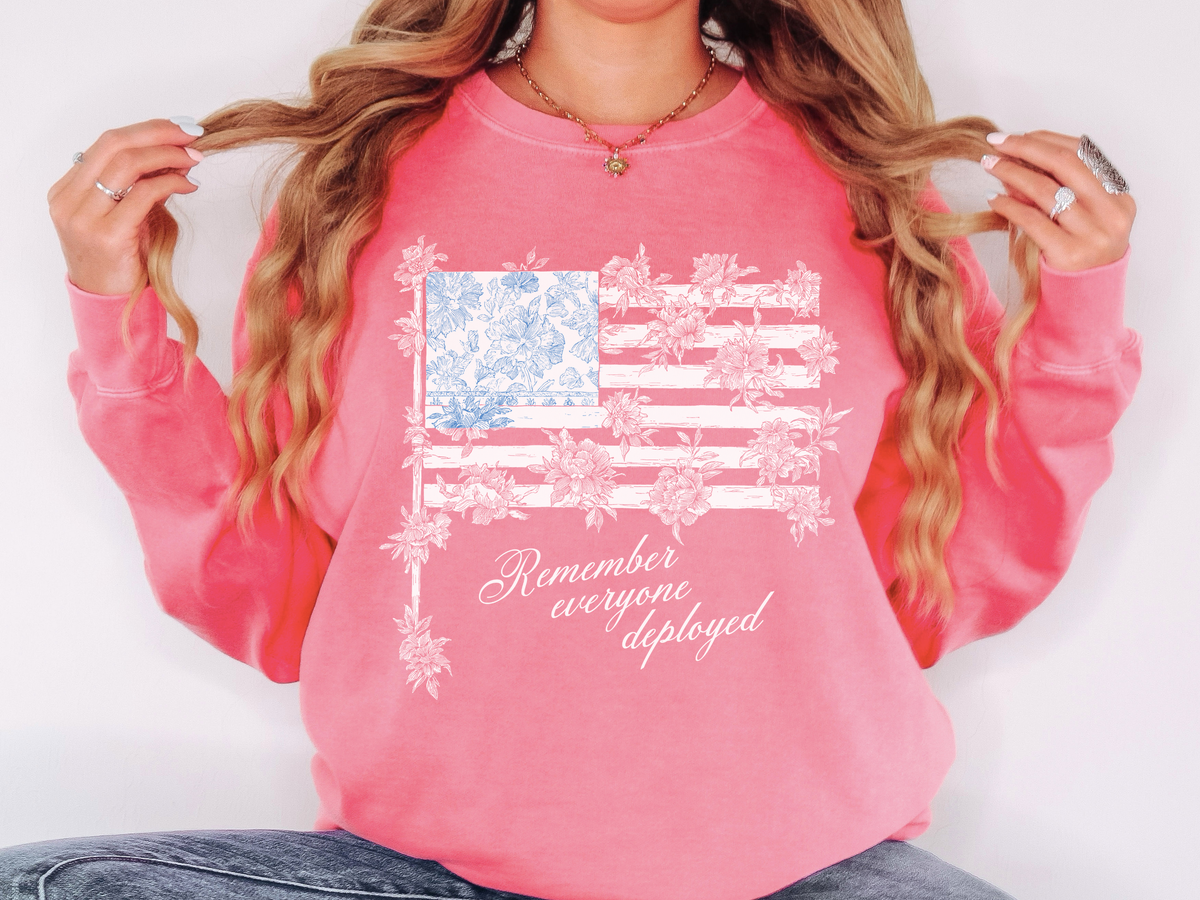 A pink crewneck sweatshirt printed with a white floral American flag with a blue toile starfield above Remember everyone deployed in script.