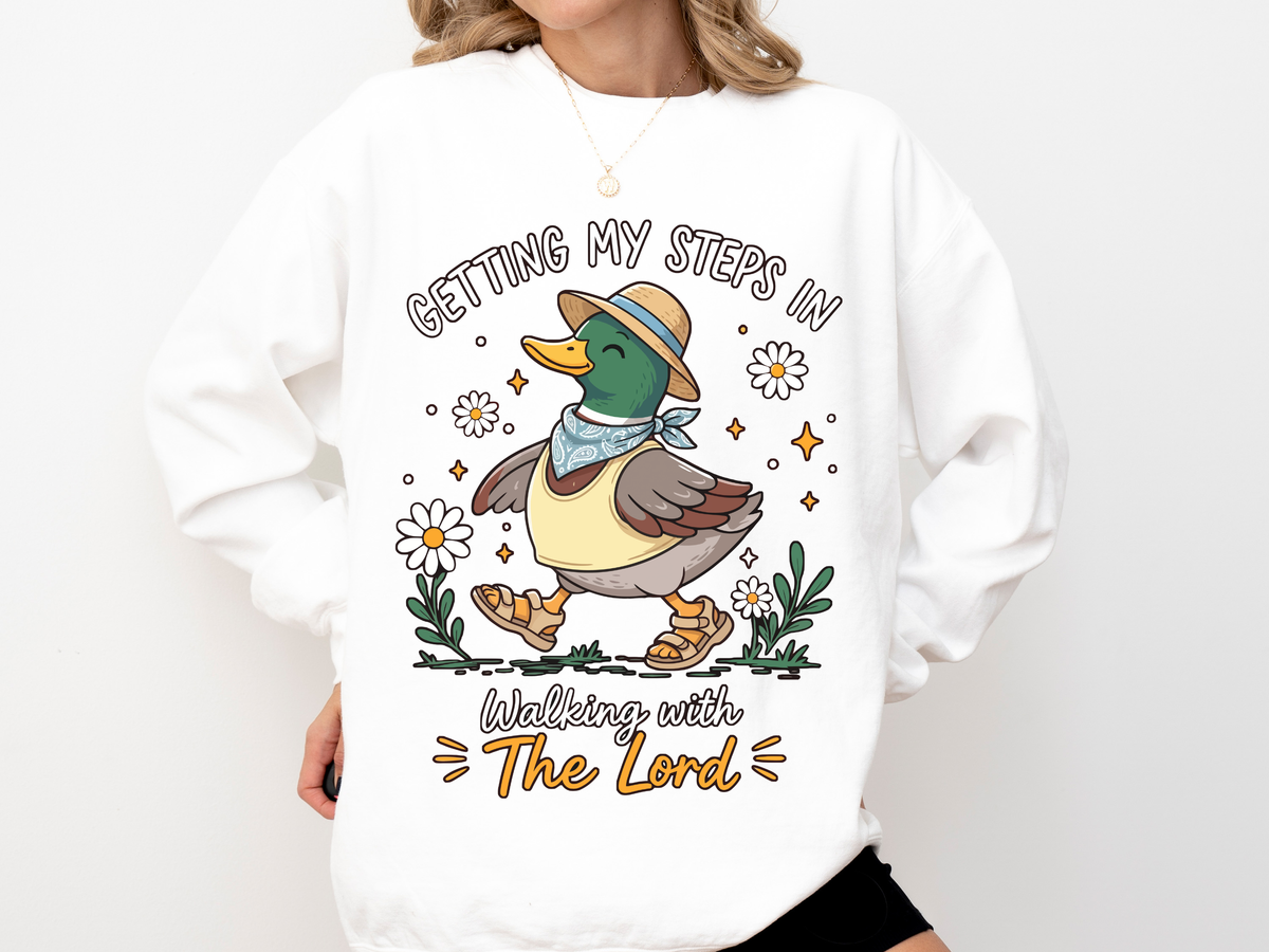 A white crewneck sweatshirt printed with a duck in a straw hat walking among daisies with the words Getting my steps in, Walking with The Lord.