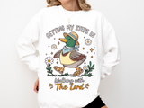 A white crewneck sweatshirt printed with a duck in a straw hat walking among daisies with the words Getting my steps in, Walking with The Lord.