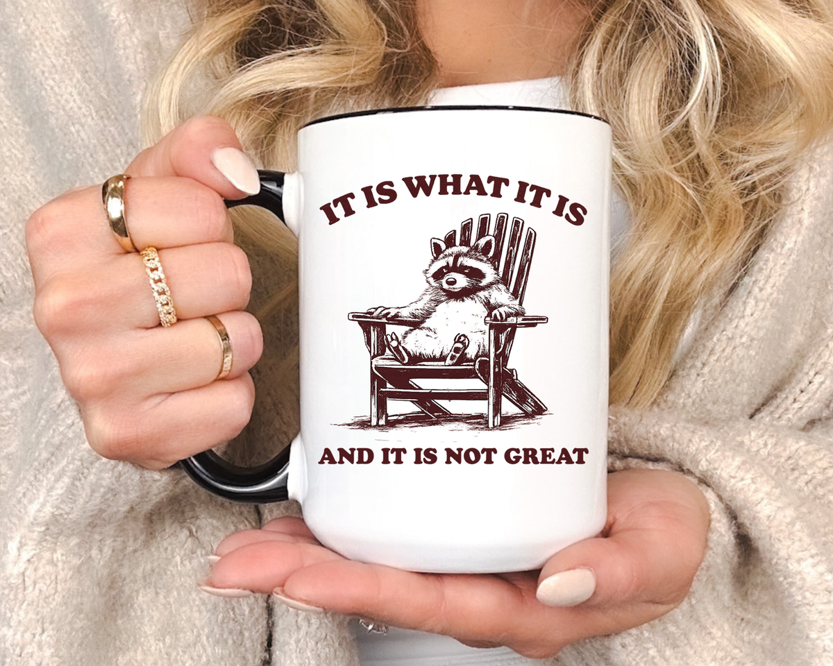 White mug with a black handle and rim, printed in dark red with a raccoon lounging back in a wooden deck chair, headed It Is What It Is and finished And It Is Not Great below.