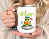 White mug with a black handle and rim, printed with a green alien sitting cross legged on a flying saucer, reading an orange book titled Being Human beside a lightbulb and a brain.