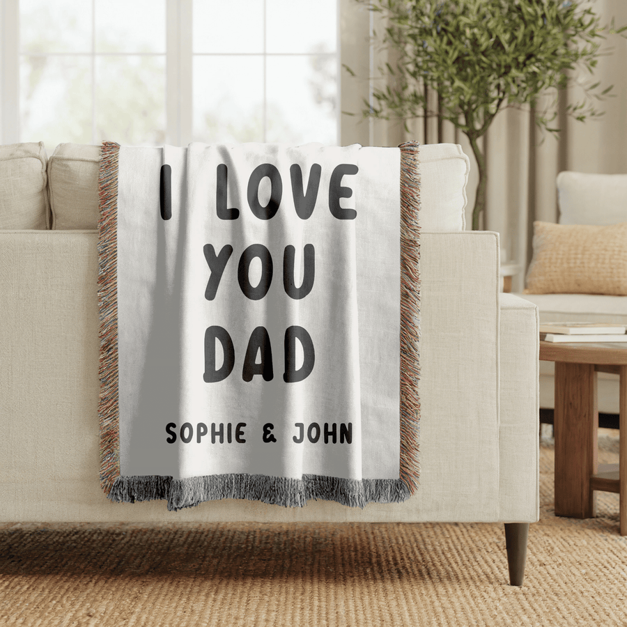 White fringed woven blanket printed with the words I Love You Dad in tall bold black capitals across three lines, with the names Sophie and John set in a row beneath them.