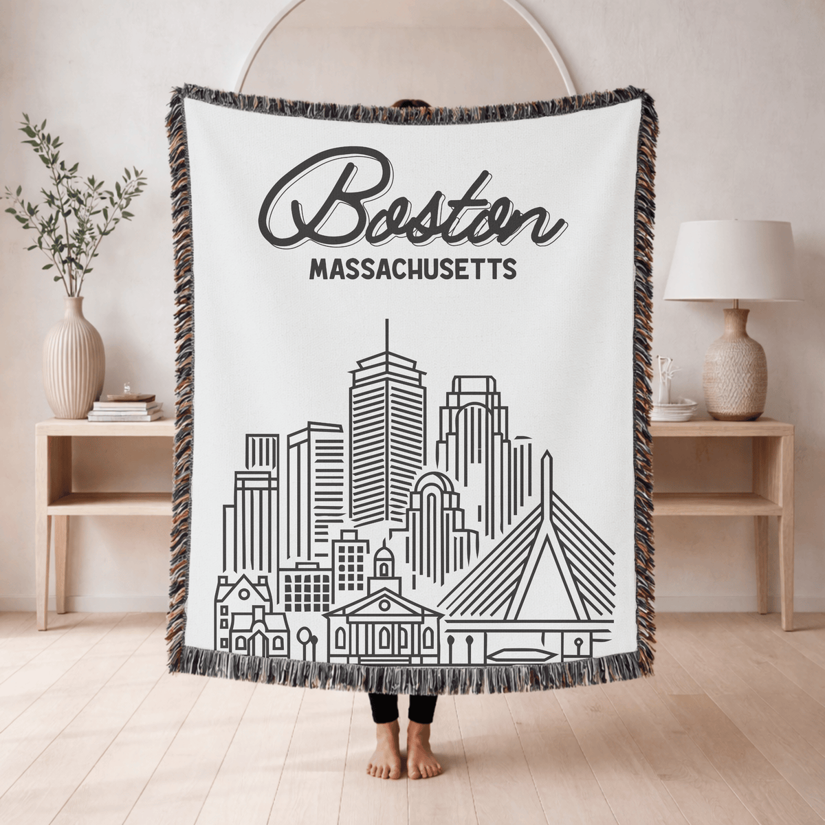 White fringed woven blanket reading Boston in bold script above the word Massachusetts, over a fine line drawing of the skyline with a domed church and a cable-stayed bridge.