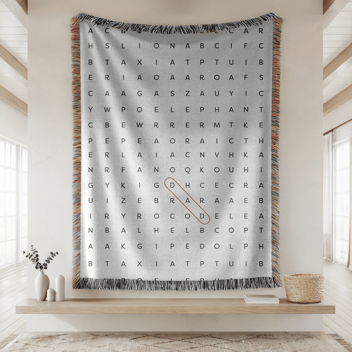 Grey fringed woven blanket printed as a large word-search grid of grey capital letters, with a single word ringed in an orange oval running diagonally through the middle.