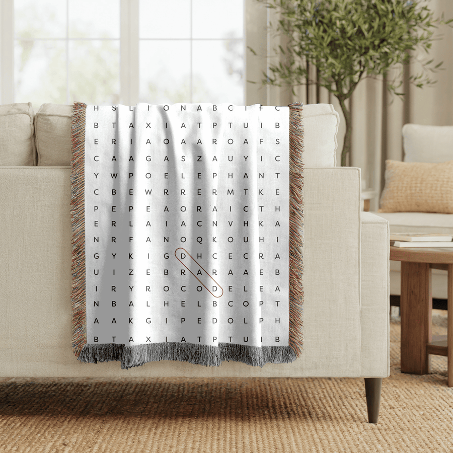 Grey fringed woven blanket printed as a large word-search grid of grey capital letters, with a single word ringed in an orange oval running diagonally through the middle.