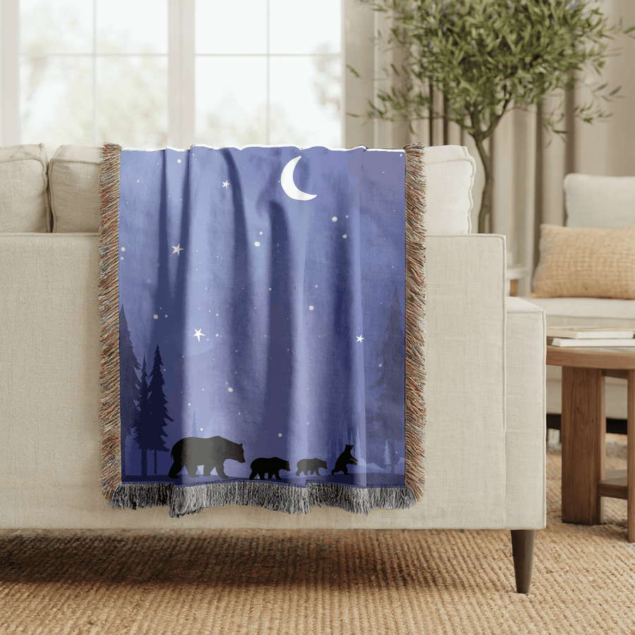 Deep blue fringed woven blanket printed with a crescent moon and stars over a pine treeline, with a mother bear and three cubs walking in silhouette along the lower edge.