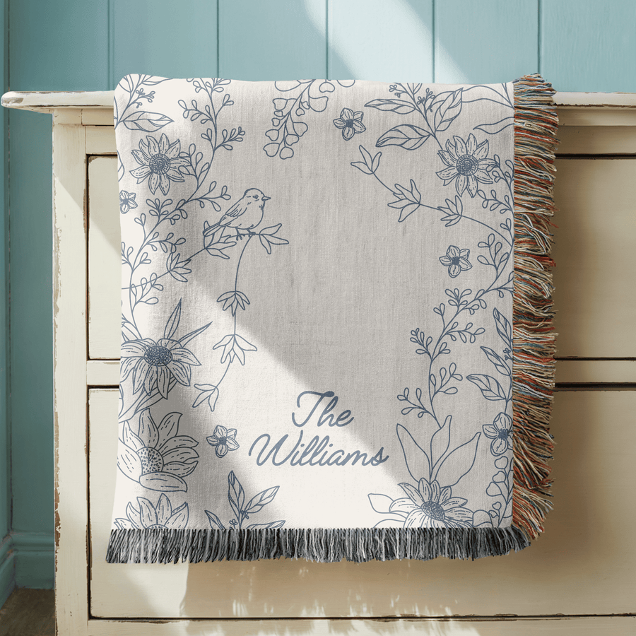 Cream fringed woven blanket printed with a blue line-drawn border of sunflowers and trailing stems with a small bird perched among them, reading The Williams in blue script.