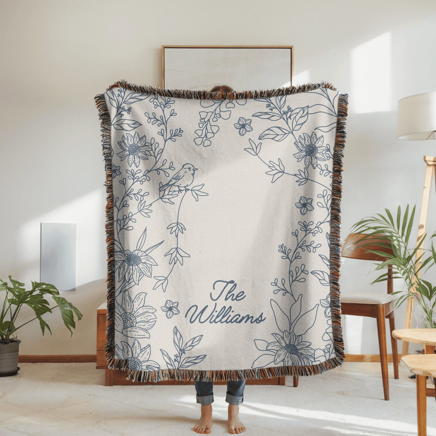 Cream fringed woven blanket printed with a blue line-drawn border of sunflowers and trailing stems with a small bird perched among them, reading The Williams in blue script.