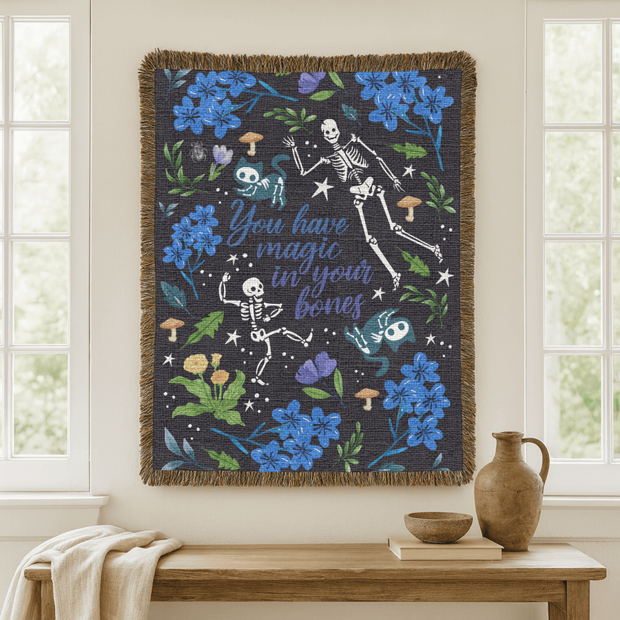 Charcoal fringed woven blanket reading You have magic in your bones in blue script, surrounded by dancing skeletons, blue flowers, toadstools, cat-faced moths and scattered stars.