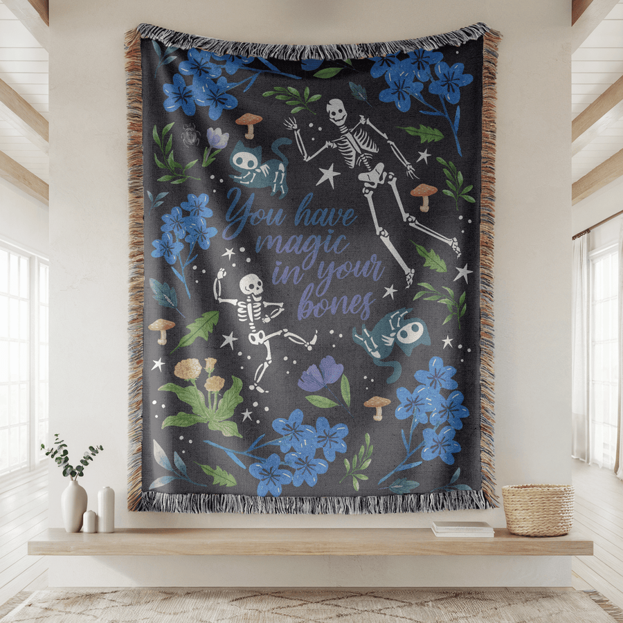 Charcoal fringed woven blanket reading You have magic in your bones in blue script, surrounded by dancing skeletons, blue flowers, toadstools, cat-faced moths and scattered stars.
